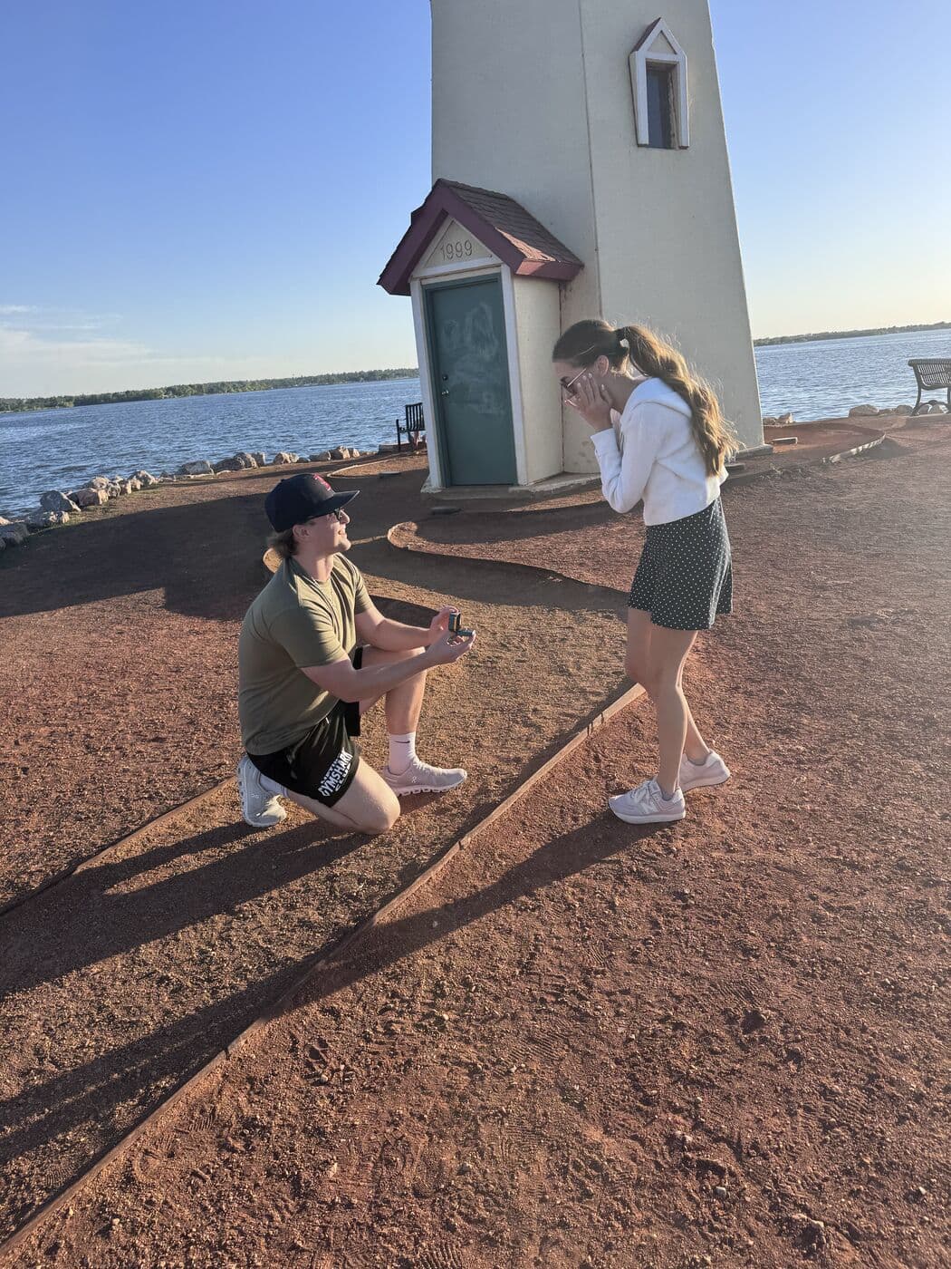 Tyler kneeling on the red dirt path by the lighthouse as Elise covers her face